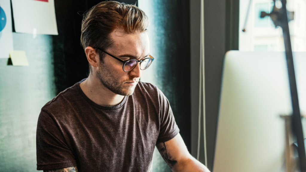 Bearded man in glasses working at a desktop computer at a wooden desk