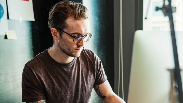 Bearded man in glasses working at a desktop computer at a wooden desk