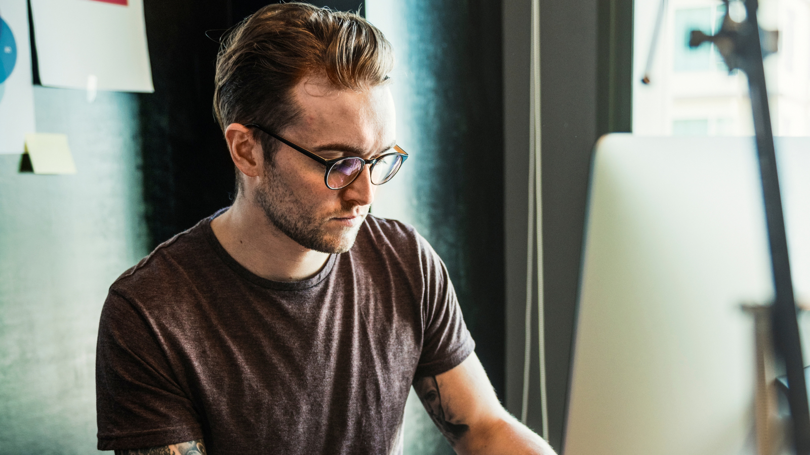 Bearded man in glasses working at a desktop computer at a wooden desk