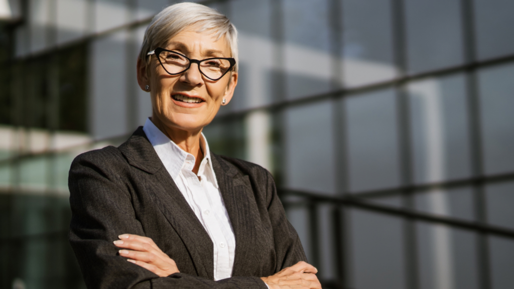 Silver-haired woman in a gray suit standing with arms crossed outside a glass building