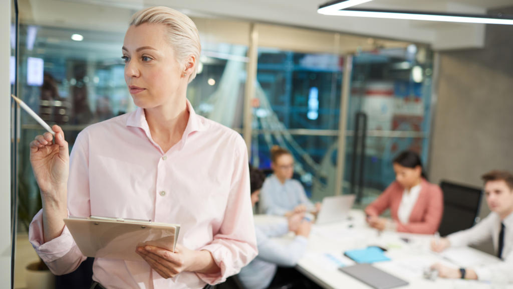 Woman in a pink shirt holding a clipboard, colleagues meeting behind her in a modern office