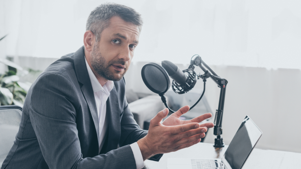 Gray-haired man in a suit gesturing while speaking into a podcast microphone