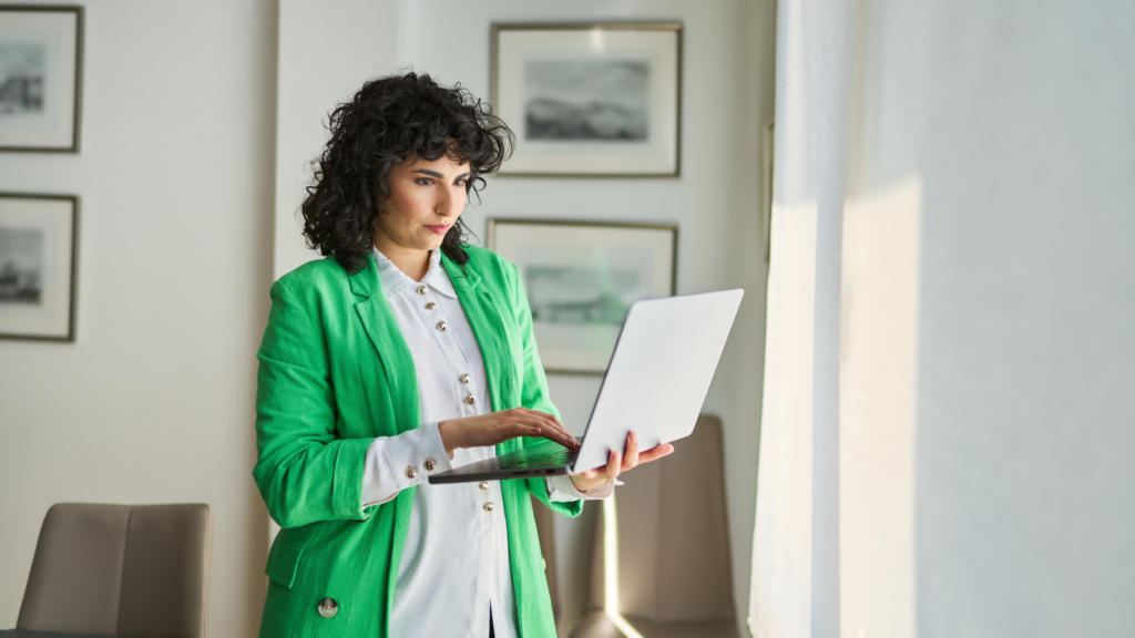 Woman in a green jacket working on a laptop by a window in a modern office