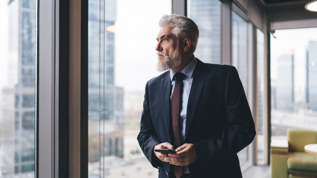 Gray-bearded executive in a suit holding a phone, looking out an office window