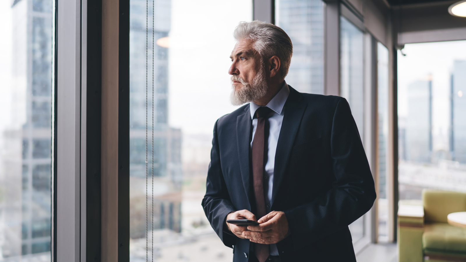 Gray-bearded executive in a suit holding a phone, looking out an office window