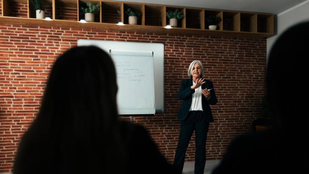 Gray-haired woman presenting to a seated audience beside a whiteboard